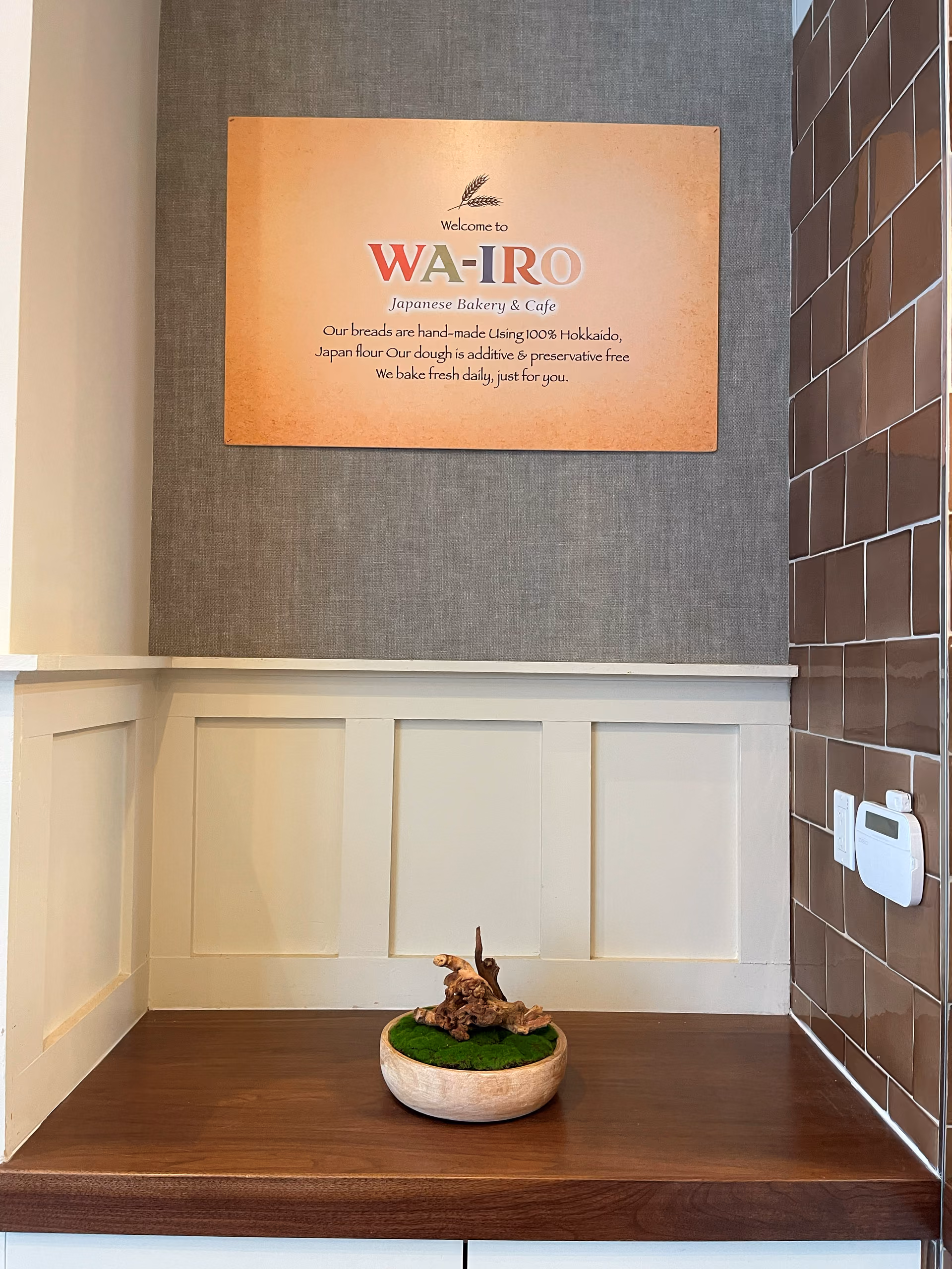 A framed welcome sign reading "Welcome to WA-IRO, Japanese Bakery & Café" above a small moss arrangement on a wood counter.