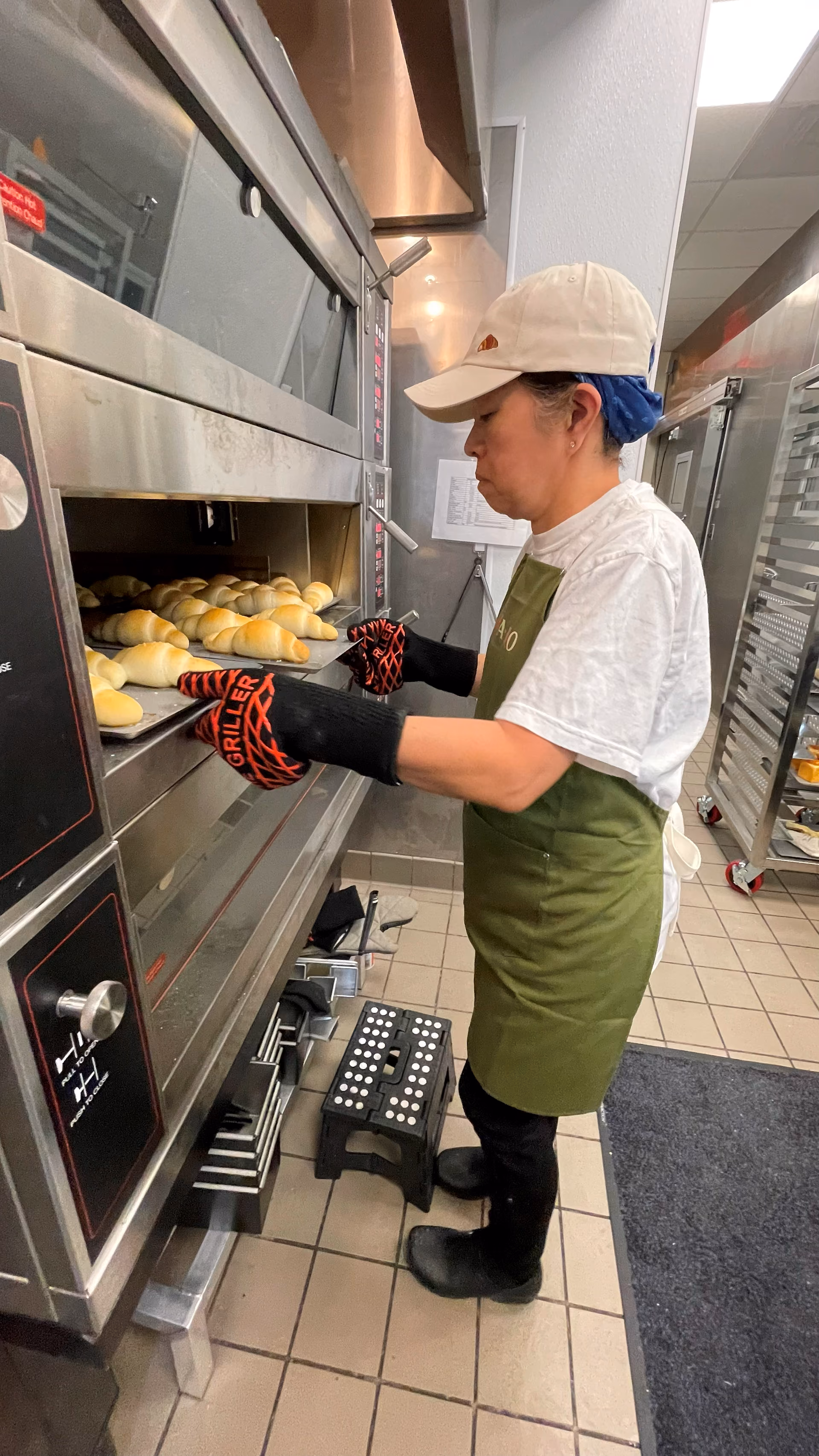 A tray of golden-baked rolls fresh from the oven, held by a staff member in the bakery kitchen.