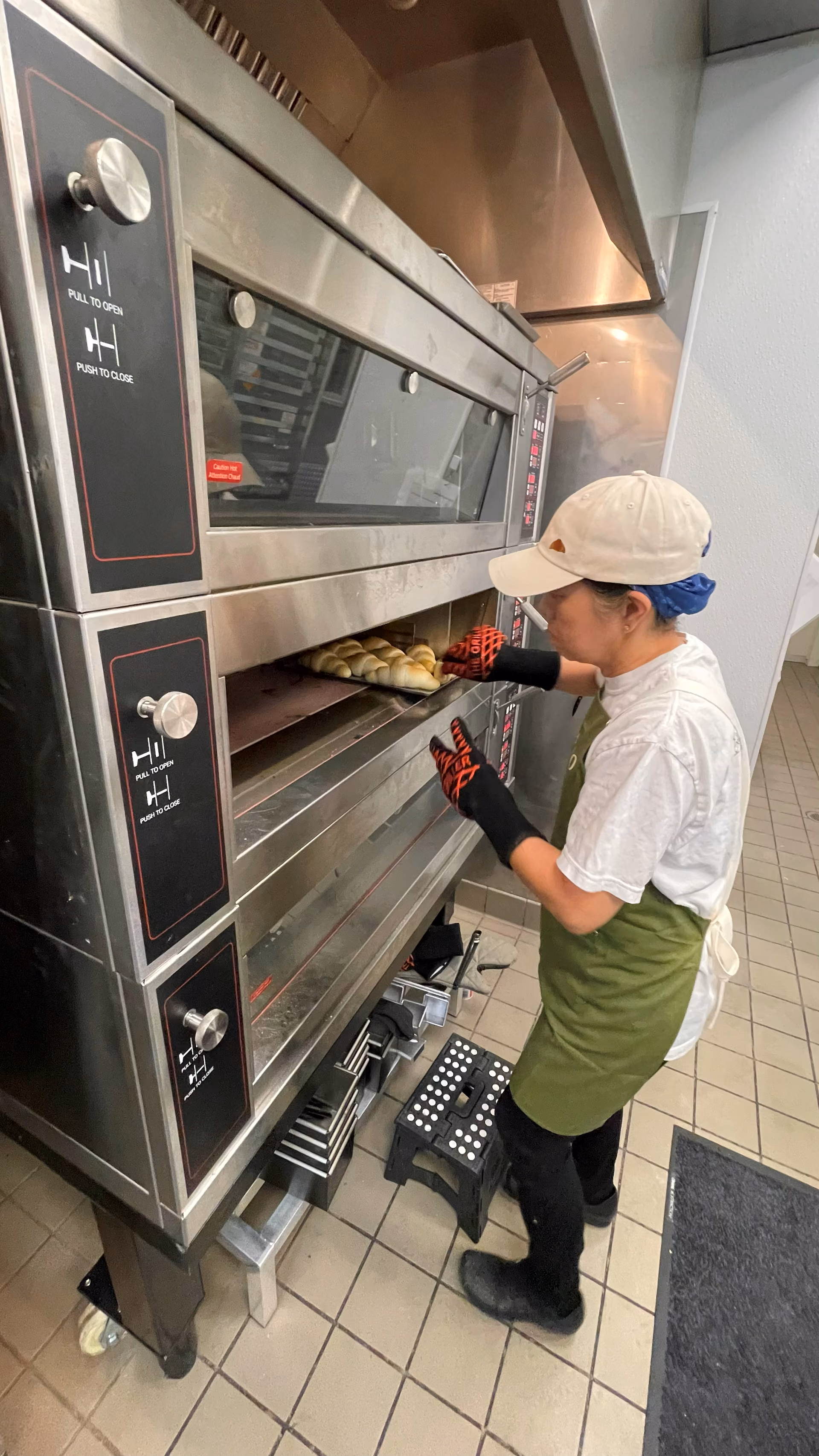 A staff member in oven mitts pulling a tray of fresh rolls from a deck oven.