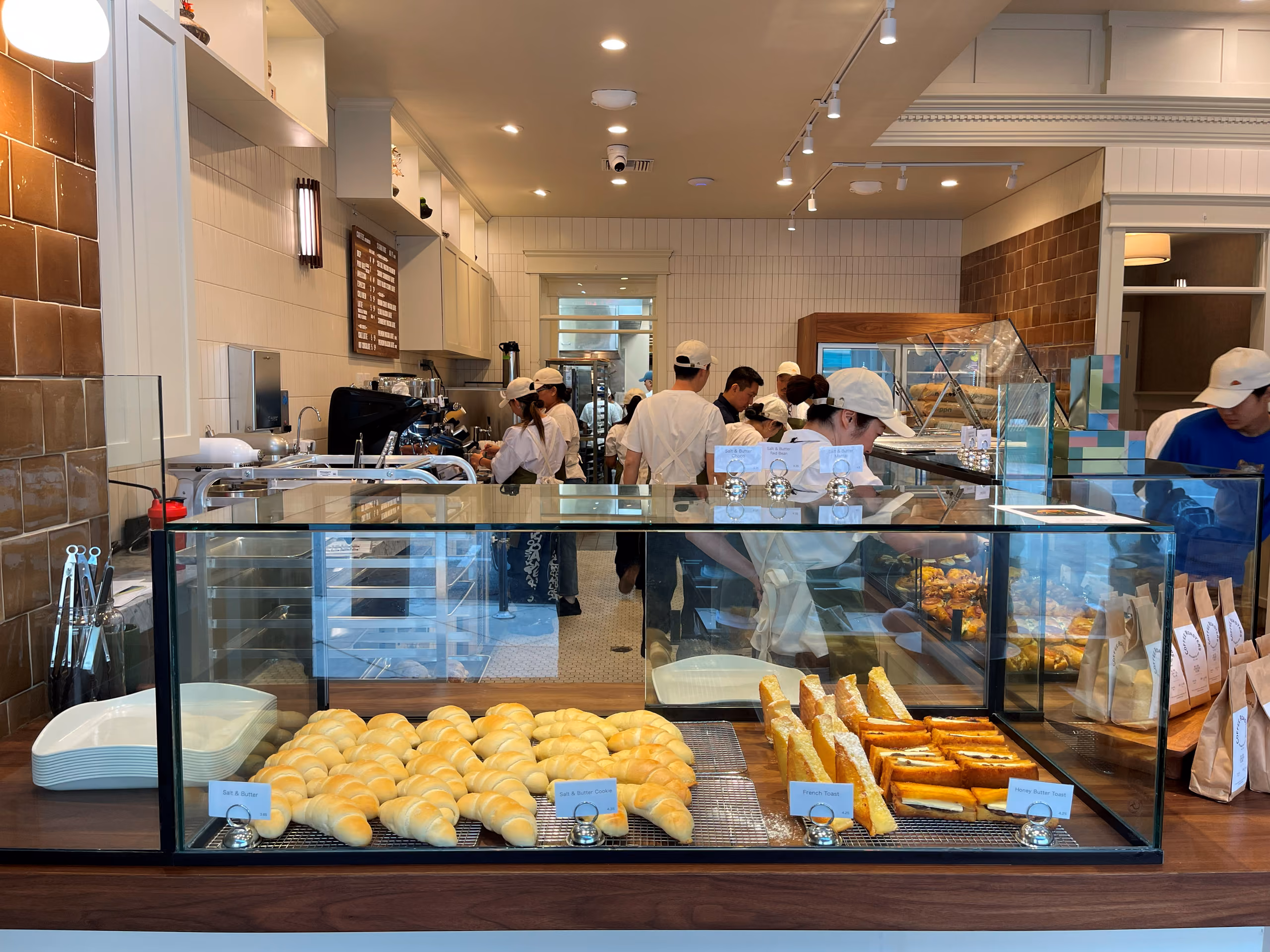 A wood-topped glass bakery case with rows of croissants and toasts, warm pendant lighting, staff at work in the background.