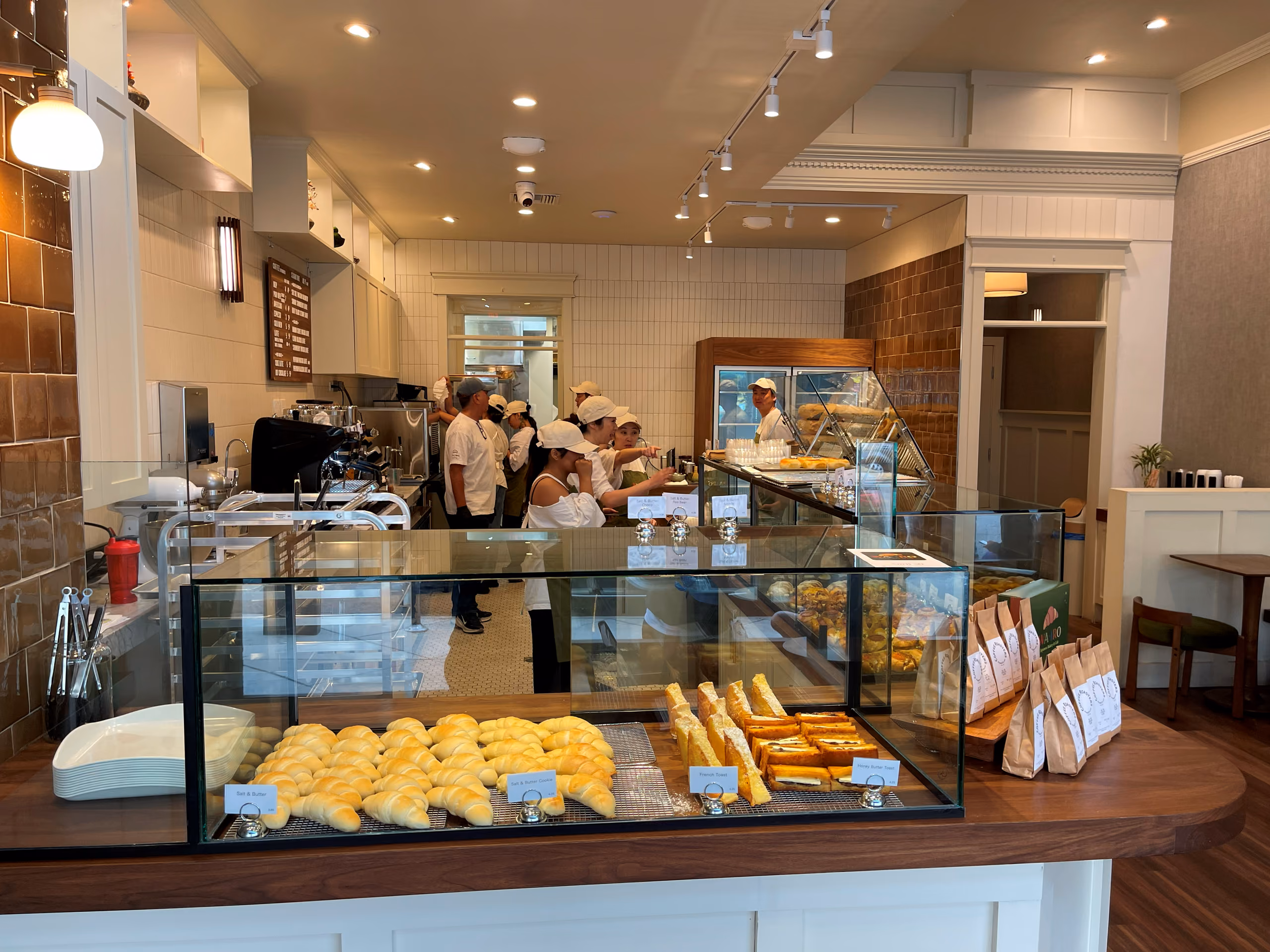 A bakery case of croissants and pastries with paper takeout bags stacked beside it, staff preparing orders behind the counter.
